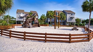 Zona arenosa de playa con valla de madera, parque infantil y casas al fondo.