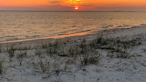 Una playa con arena y vegetación escasa al frente, y una puesta de sol sobre el agua.
