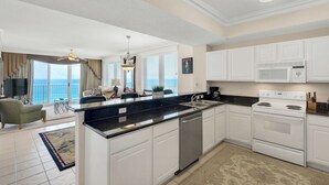 A modern kitchen with white cabinets, a black countertop, and stainless steel appliances. There is a dining area with a view of the ocean. - Captain`s Hideaway - Summerwind 905E (Navarre)