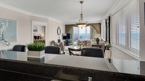 A spacious living area with a dining table, comfortable seating, a television, and a view of the ocean through large windows. - Captain`s Hideaway - Summerwind 905E (Navarre)