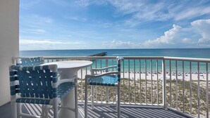 Un balcon avec deux chaises bleues et une petite table, donnant sur une plage avec une jetée et des personnes que l’on aperçoit au loin.
