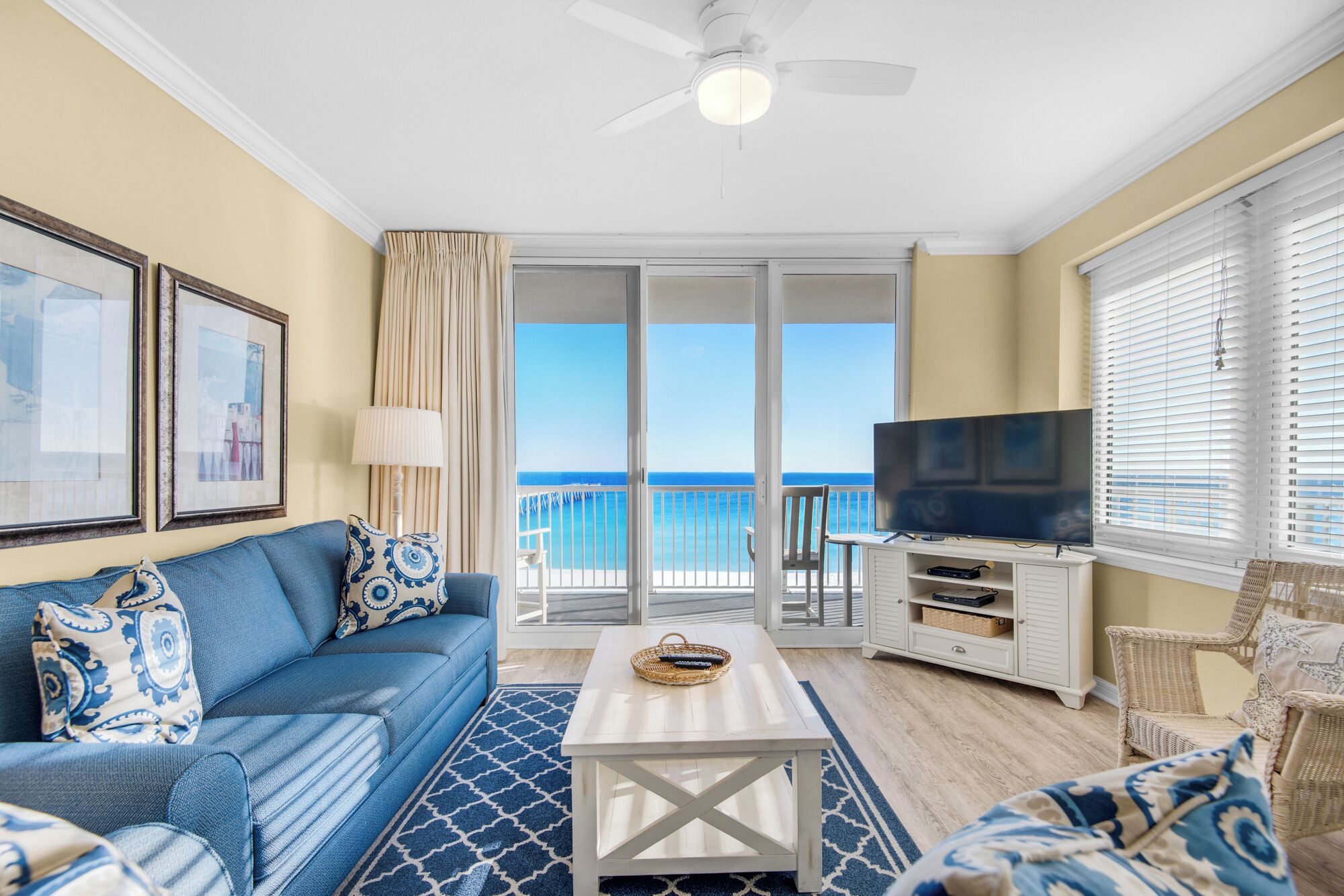 A living room with a blue sofa, a white coffee table, a TV, and a balcony with a view of the ocean.