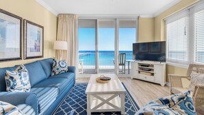 Un salon avec un canapé bleu, une table basse blanche, un téléviseur et un balcon offrant une vue sur l’océan.