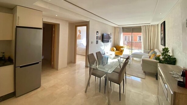Una cocina moderna con refrigerador empotrado, un comedor con mesa y sillas de vidrio, y una sala de estar con sofá y televisor.