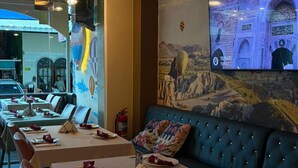 Ein Restaurant mit langen Tischen, einer blauen Sitzgelegenheit mit Polstern und einem Wandbild einer Landschaft.