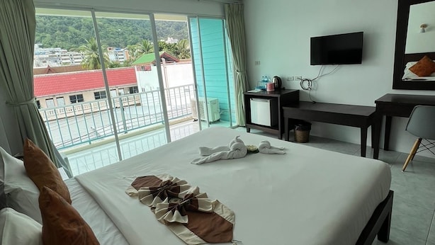 Ein Hotelzimmer mit einem großen Bett, einem Schreibtisch mit Stuhl, einem Fernseher und einem Balkon mit Blick auf Gebäude und Grünflächen.