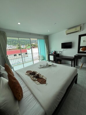 A hotel room with a large bed, a desk with a chair, a television, and a balcony with a view of buildings and greenery. - Kiwi Boutique Hotel (Patong)