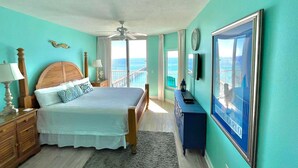 A bedroom with a bed, a nightstand, a dresser, a ceiling fan, a window with a view of the ocean, and a framed picture on the wall.