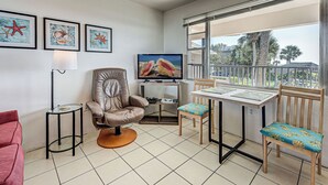 A living room with a TV, a red chair, a brown leather chair, a small table, and a balcony with a view. - Directly on the Gulf - Unit 14 - 1 Bedroom - Second Floor - Sleeps up to 5 (Venice)