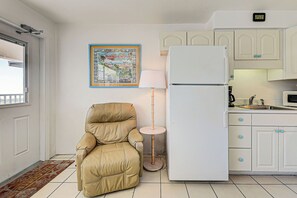 A kitchen with white cabinets, a white refrigerator, a beige armchair, a floor lamp, and a framed map on the wall. - Directly on the Gulf - Unit 14 - 1 Bedroom - Second Floor - Sleeps up to 5 (Venice)