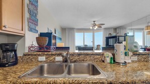A kitchen with granite countertops, stainless steel sinks, a coffee maker, and a view of the ocean through large windows.