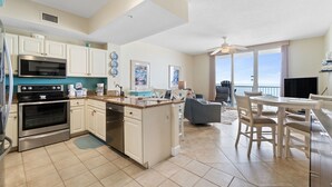 A modern kitchen with white cabinets, stainless steel appliances, and a central island. There is a dining area with a table and chairs, and a balcony with a view.