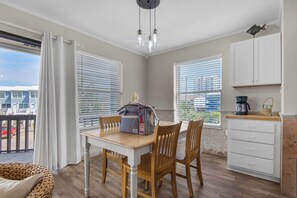A dining area with a table and chairs, a wicker chair, a bag, and a kitchen area with cabinets and a coffee maker.
