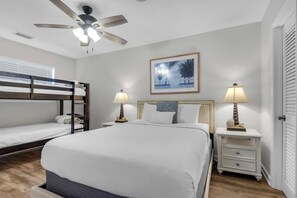 A hotel room with a bunk bed, a single bed, a ceiling fan, two bedside tables with lamps, a framed picture on the wall, and a door with blinds.