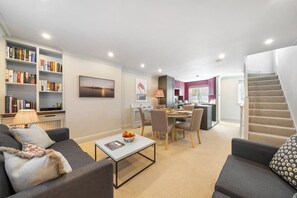 A modern living room with a sofa, coffee table, and bookshelves. There is a dining area with chairs and a table, and a kitchen in the background.