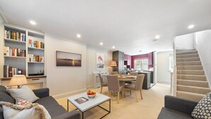 A modern living room with a sofa, coffee table, and bookshelves. There is a dining area with chairs and a table, and a kitchen in the background. - Modern 3-floor open plan townhouse with patio (London)