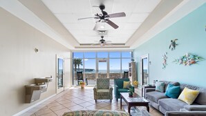 Un vaste espace de vie comprenant un ventilateur de plafond, un canapé, une table basse et un fauteuil. Une fenêtre offre une vue sur la plage et les palmiers.