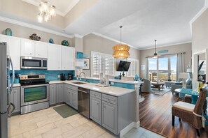 A modern kitchen with stainless steel appliances, a dining area with wicker furniture, and a view of the beach. - Stow Away - Summerwind 101C (Navarre)