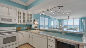 A kitchen with white cabinets, a stove, and a dishwasher. There are multiple windows with blinds, a ceiling fan, and a sign that says "Go Surfing!". - Sanctuary - Summerwind 501C (Navarre)