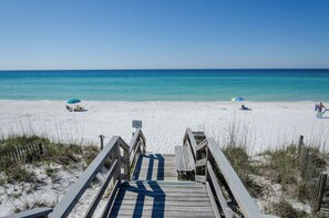 Une promenade en bois mène à une plage où l’eau est d’un bleu cristallin, et où l’on aperçoit quelques parasols de plage.