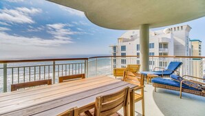 A deck with a wooden table and chairs, a small table with a chair, and a lounge chair with a blue cushion. The deck overlooks the ocean and a building with multiple balconies. - Pelican Penthouse - Beach Colony West PH 17D (Navarre)