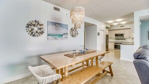 A dining area with a wooden table, white wicker chair, and a bench. There is a chandelier above the table and a beach-themed painting on the wall. - Pelican Penthouse - Beach Colony West PH 17D (Navarre)