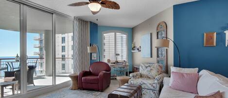 A room with a large window, a ceiling fan, a desk, a red chair, a white sofa, and a suitcase.