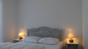 A neatly made bed with white bedding and pillows, flanked by two bedside tables with lamps, in a simple room with plain walls.