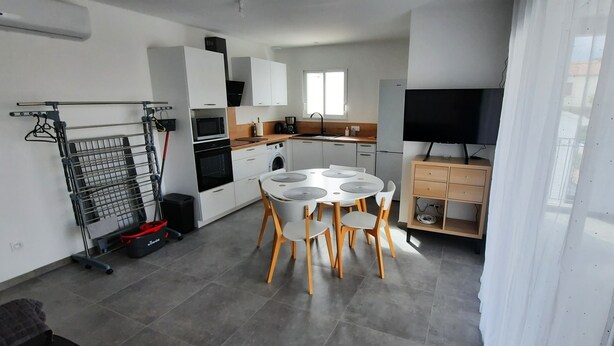A modern kitchen with white cabinets, a dining area with a round table and chairs, a washing machine, and a TV on a wooden cabinet.