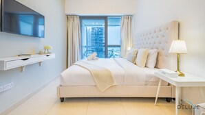 A modern hotel room with a large bed, a TV mounted on the wall, a desk with a lamp, and a view of a cityscape through the window. - Charming Studio in Saba 3, Jumeirah Lake Tower by Deluxe Holiday Homes (Dubai)