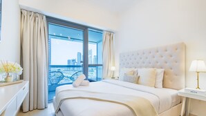 A hotel room with a large bed, a nightstand with a lamp, and a view of a cityscape through a large window. - Charming Studio in Saba 3, Jumeirah Lake Tower by Deluxe Holiday Homes (Dubai)