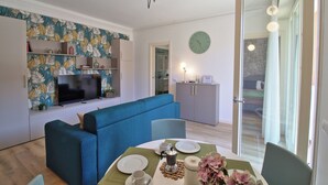 Una sala moderna con un sofá azul, una mesa de comedor con platos y tazas, un televisor sobre un mueble blanco y un reloj de pared.