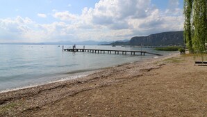 Una playa con un muelle de madera que se adentra en aguas tranquilas, bordeada por una orilla pedregosa y montañas a lo lejos.
