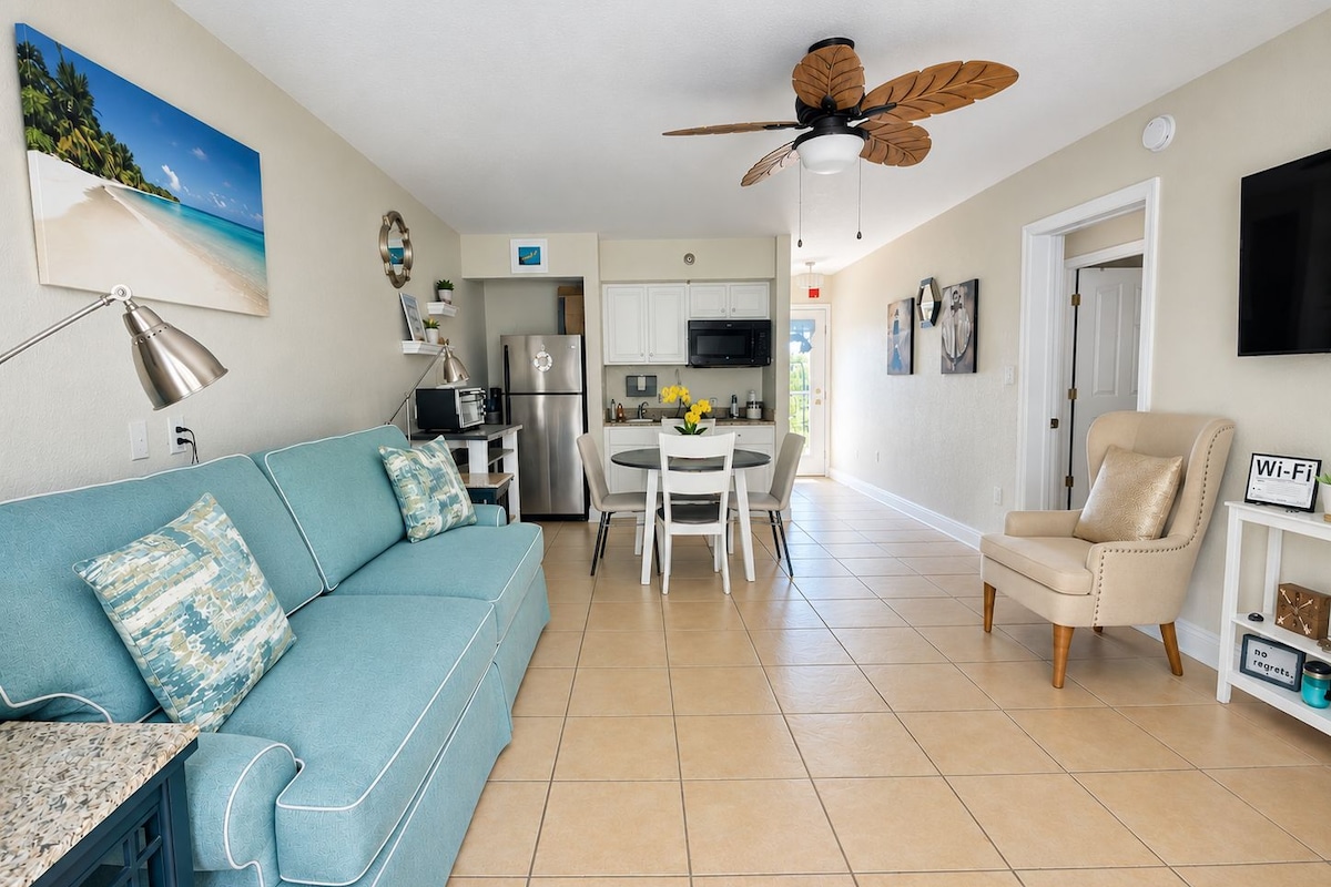 A modern living room with a blue sofa, a dining table, and a kitchen area with a refrigerator and microwave.