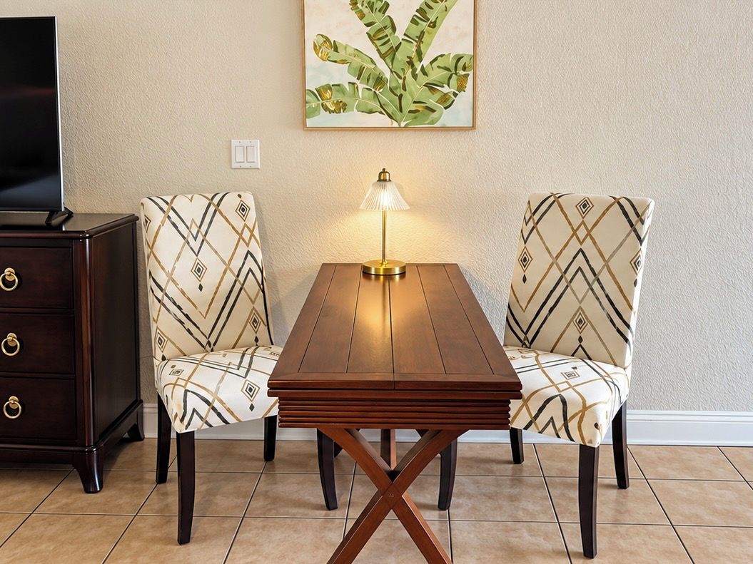 A dining area with a wooden table, two upholstered chairs, a lamp, and a television on a cabinet.