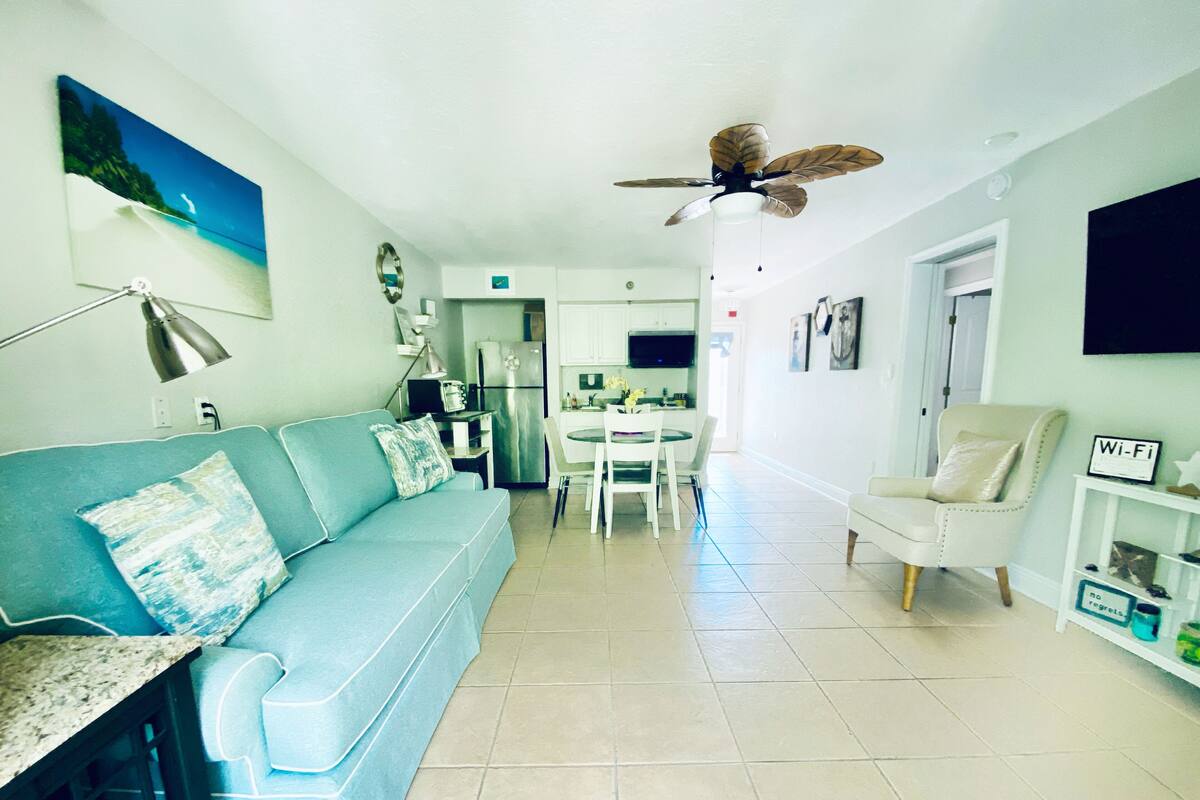 Un salon moderne avec un canapé bleu, un fauteuil blanc, un coin repas avec une table et des chaises, une cuisine équipée et un ventilateur de plafond.