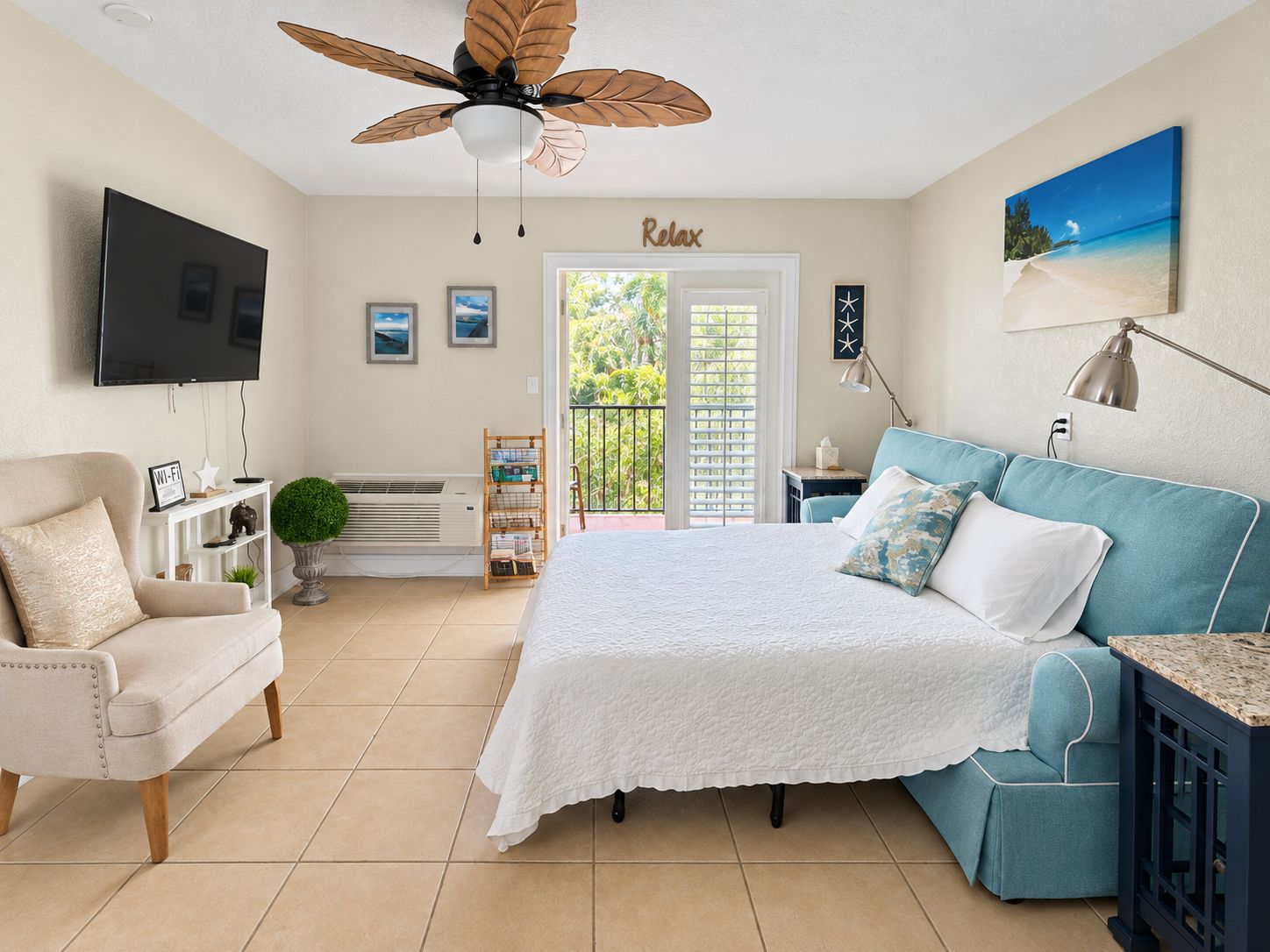 A bedroom with a bed, a chair, a TV, a ceiling fan, a balcony door, and a painting.