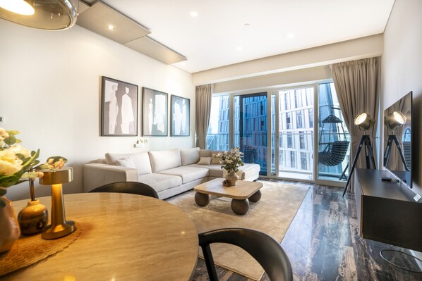 Una sala de estar moderna con sofá, mesa de centro y mesa de comedor. Cuenta con ventanas grandes con cortinas, un televisor y obras de arte en las paredes.