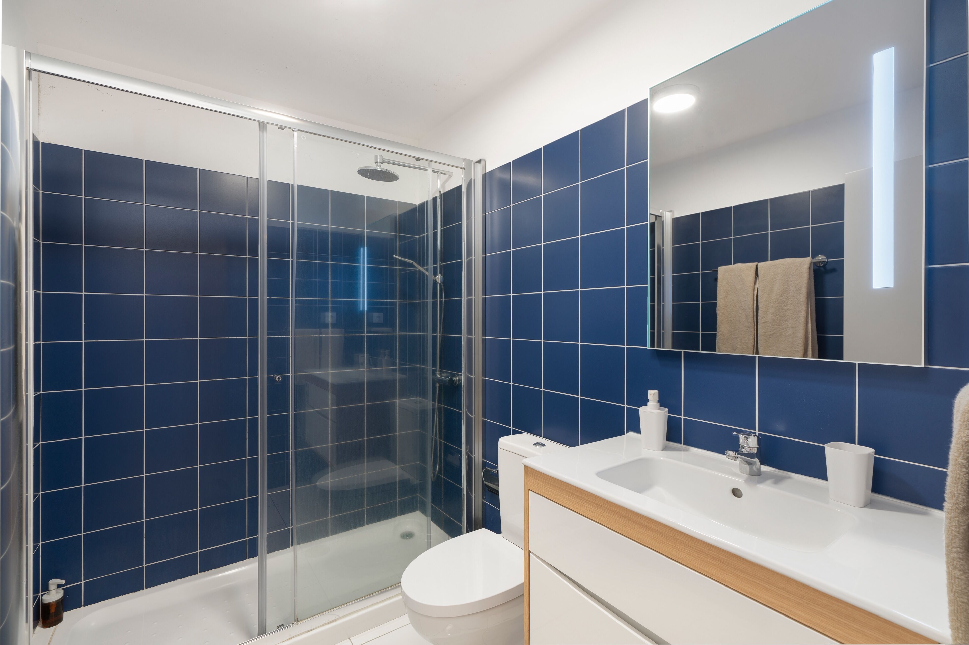 A modern bathroom with blue tiles, a glass shower enclosure, a white toilet, and a sink with a mirror.