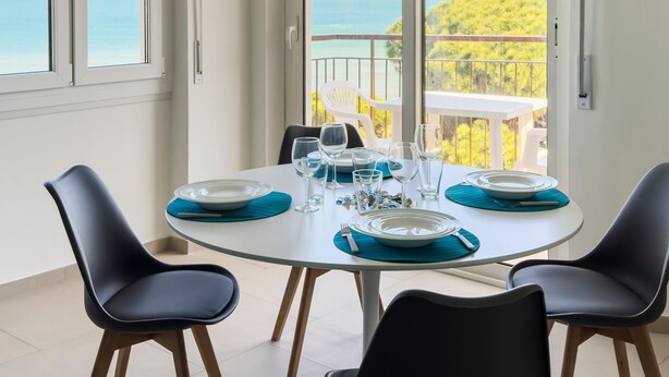 Een eetgedeelte met een ronde tafel voor vier personen, grote ramen met uitzicht op zee en groen, en een balkon met buitenmeubilair.
