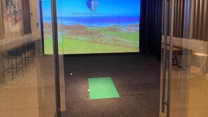 Quarto com uma tela grande exibindo 'HIGH DEFINITION GOLF' e um tapete verde no chão.