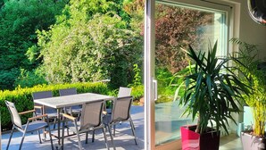 A balcony with a table and chairs, a glass door, and a view of a lush garden. - Schöne 120 qm Ferienwohnung + 50 qm Sonnenterrasse im Grünen-stockelsdorf/lübeck (Stockelsdorf)