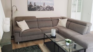 A modern living room with a brown sectional sofa, a glass coffee table, a floor lamp, and a painting of a rocky landscape. - Schöne 120 qm Ferienwohnung + 50 qm Sonnenterrasse im Grünen-stockelsdorf/lübeck (Stockelsdorf)