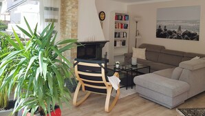 A living room with a fireplace, a sofa, a bookshelf, and a large window. - Schöne 120 qm Ferienwohnung + 50 qm Sonnenterrasse im Grünen-stockelsdorf/lübeck (Stockelsdorf)