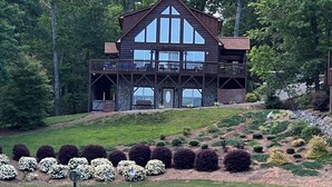 A large, two-story wooden lodge with multiple balconies and large windows, surrounded by lush greenery and a well-maintained lawn.