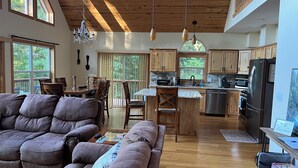 A spacious living room with a large brown sectional sofa, a wooden dining table, and a kitchen area with wooden cabinets and stainless steel appliances.