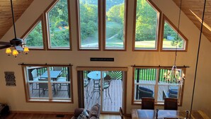 A spacious room with a high ceiling, large windows offering views of a lush landscape, a dining area with a table and chairs, and a cozy seating area with a couch.