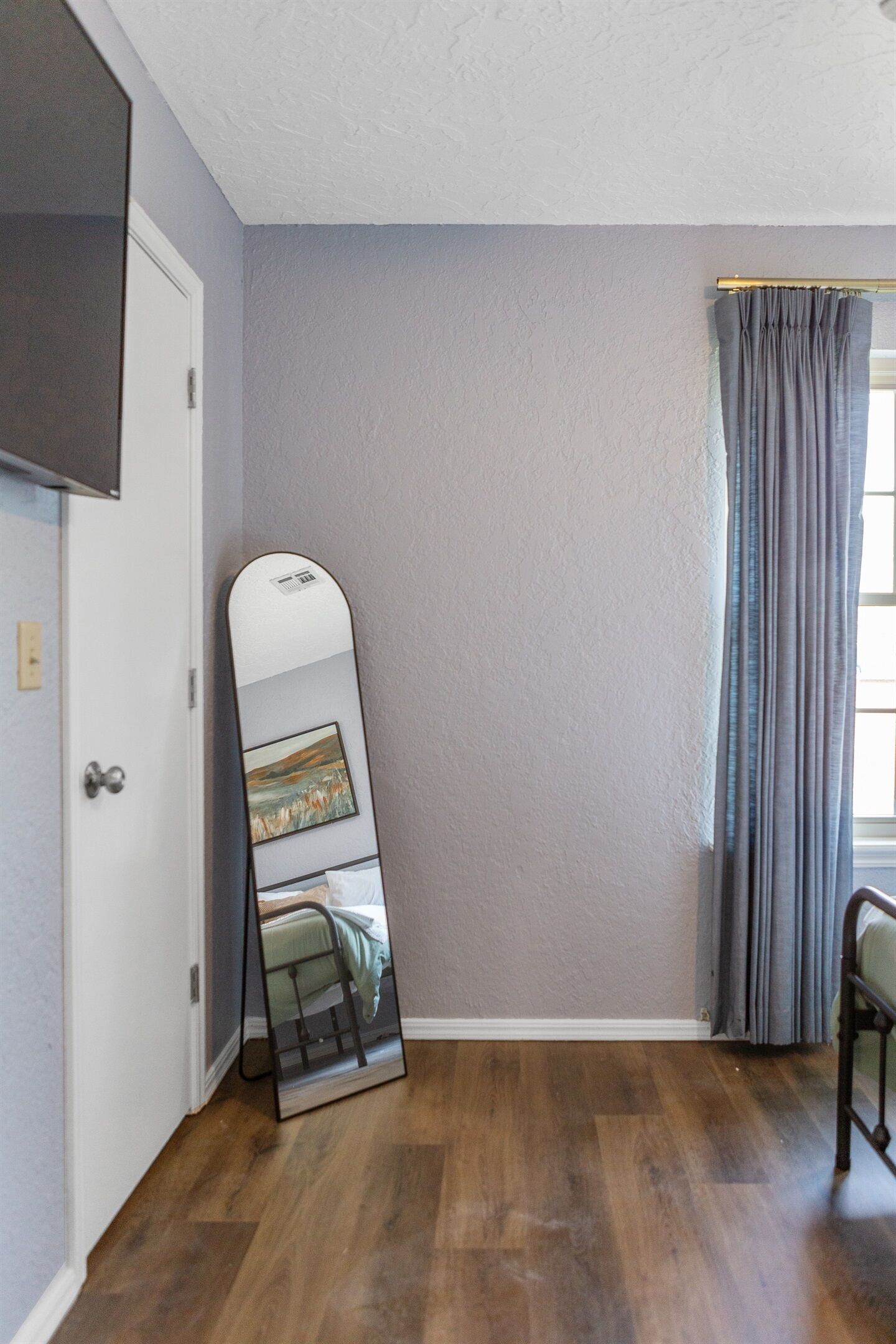 A room with a wooden floor, a white door, a large mirror leaning against the wall, and a window with grey curtains.