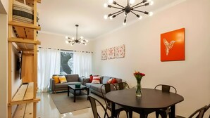Een moderne woonkamer met een bank, salontafel en eettafel. Er is een trap met houten leuning, een plafondlamp en een kunstwerk aan de muur.
