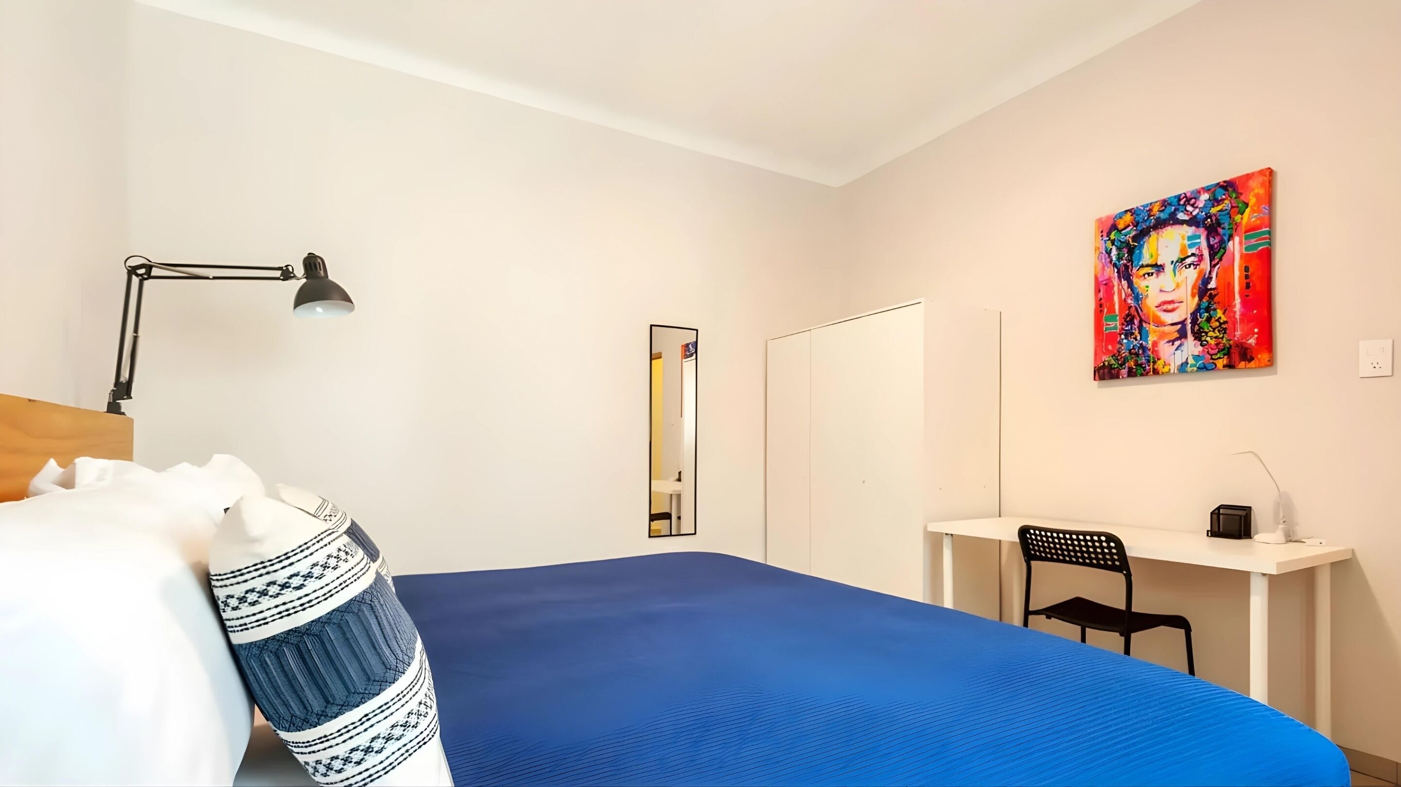 Een slaapkamer met een bed, een bureau, een stoel, een lamp en een schilderij.